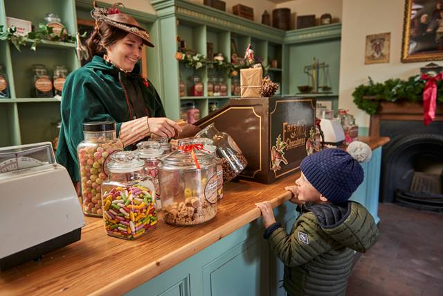 child at blue counter with wooden worktop with jars of sweets sitting on it, looking up as woman in Victorian clothing both smiling