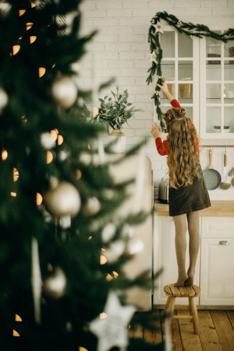 Picture of young girl with light brown hair hanging a green Christmas garland aroudn the top of kitchen cabinets with a Christmas tree decorated with white decorations out of focus in front and to the left of her.
